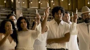 A group of men and women, all clad in white, stand inside a barn golding lit white candles aloft in their hands.