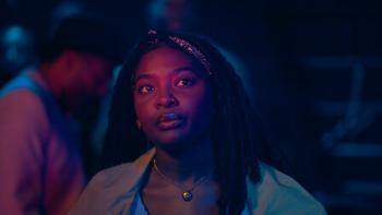 Déja J. Bowens stands in a crowd, watching a performance on stage. She is case in blue and pink light.