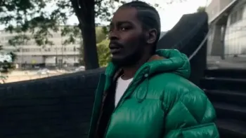 Malcolm Kamulete wears a green puffer jacket and white t-shirt. She stands on the steps outside of a building.