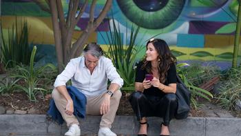 Chivalry TV show screenshot 3: Cameron and Bobby sit on a curb, a colorful mural behind them. Cameron bows his head, Bobby holds her phone and smiles