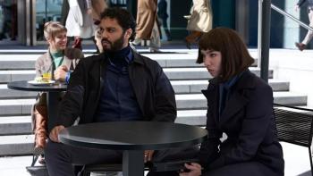 (From R-L) Marouane Zotti and Matilda De Angelis sit at a table. Angelis holds a pistol between her legs, aiming it forward.