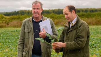 Two aging white men in overcoats stand in a field. One holds a freshly picked piece of produce while the other clutches papers
