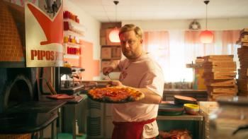 Crap Happens TV show screenshot 1: A man in a pizzeria holds a pizza near an oven, with pizza boxes stacked behind him.