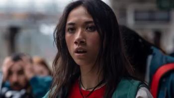 Zoë Boe stands outside amongst a crowd of people. She wears a red shirt, black necklace, and green hooded jacket.