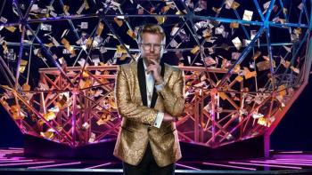 The Crystal Maze TV show: Host Adam Conover standing in front of the Crystal Dome, with right arm across body, left hand cupping his chin.