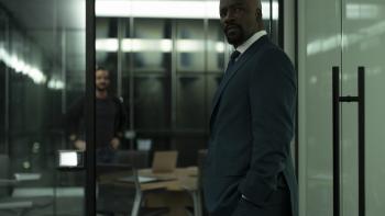 Cupertino TV show screenshot 3: Mike Colter stands in the doorway of a glass office. He wears a dark blue suit, dark tie, and white collared shirt.