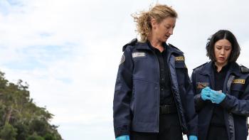 Kate Box as Sergeant Collins looks down as Nina Oyama as Constable Mutsuda (R) writes on a notepad.