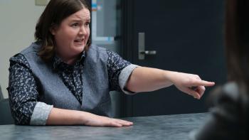 DMV TV show screenshot 2: Molly Kearney points across a table. They wear a flower printed button-up shirt with sleeves cuffed, and a dark gray vest.