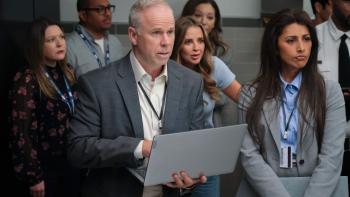 DMV TV show screenshot 3: (L-R) Gerry Dee and Reshma Shetty stand in a crowded bathroom. Dee holds a silver laptop.