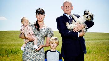 Doc Martin TV show: A man in a suit holding a dog stands next to a child and a woman holding a baby in a field.