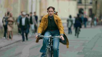 Down Cemetery Road screenshot 1: Ruth Wilson rides a bicycle down a crowded street. She wears a scarf, a yellow coat, green shirt, and blue jeans.