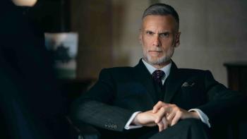 Down Cemetery Road screenshot 3: Darren Boyd sits cross legged in a leather chair. He wears a dark suit, burgundy patterned tie, and a pocket square.