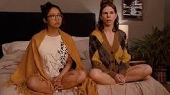 An Asian woman and a White woman sit on the bed side by side in their pajamas
