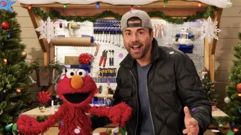 Elmo & Mark Rober's Merry Giftmas TV: Elmo and Mark Rober with their mouths open in awe while confetti falls from the sky in their workshop.