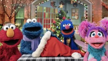 Elmo & Mark Rober's Merry Giftmas TV: Elmo, Cookie Monster, Grover and Abby stare at the camera with a santa hat laying on a table between them.