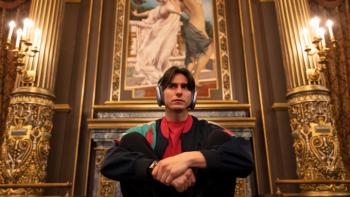 Étoile TV show screenshot 3: Tobias sits in an elegantly appointed room with elaborate carved walls, wearing headphones and making a disgusted face. 