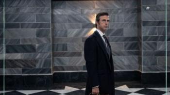 Kim Rossi Stuart stands in the safe room, wearing a long coat and holding a briefcase.