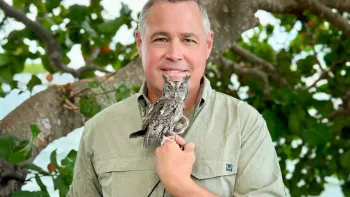 Jeff Corwin, a white, middle aged man, holds a small owl near his face while smiling at the camera and standing outdoors in front of a tree.