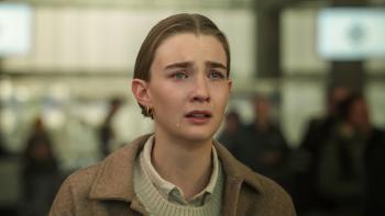 A Danish teen wearing a coat and layered clothing, with center-parted dirty blond hair and double pierced earrings cries and looks off in the distance