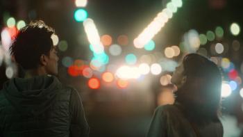 First Love TV show: A Japanese man and woman are seen standing on a street corner at nighttime, in front of them a bunch traffic lights, and car light