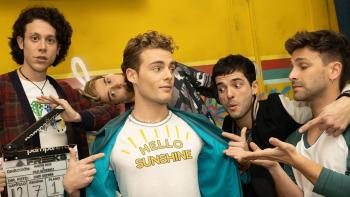 Freeks TV show screenshot 1: Three band members stand admiring their bandmates t-shirt, while another stares ahead holding a clapboard; yellow background. 