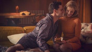 Barbara sits on a couch smiling, as her bathrobe-clad costar Clive leans toward her. Both are smiling