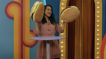 Game Changer TV show screenshot 2: Rekha Shankar wears a coral-colored dress and golds up her hands, which are covered with big plushy hamburgers