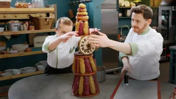 The Great British Baking Show the Professionals: a White female baker and White male baker assemble a clock tower made out of cookies