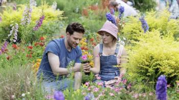 Grosse Pointe Garden Society TV show screenshot 1: Ben Rappaport and AnnaSophia Robb sit. Robb wears denim overalls, and a pink shirt and hat.