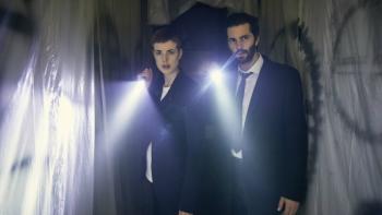 Hard Sun Series: A man and a woman, both in dark business suits, each shine their flashlights on something off-camera in front of a creepy background covered with clear sheeting and graffiti. 
