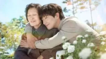 A young asian man hugs an older woman outside in a flower field. Light bathes them softly as they close their eyes in happiness.