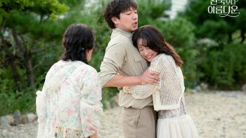 A young man in khaki pants and matching shirt is embraced by a young woman but the man is trying to pull away. An older woman looks on and watches.