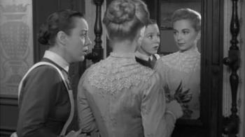 A woman with blonde hair and a white dress stands in front of a mirror as her maid stands nearby, appearing to be speaking to her urgently.