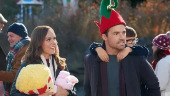 A caucasian woman with brown hair holds stuffed prizes from an outdoor fair while a brunette male wearing a green and white elf hat carries a child  
