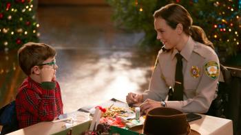 A white woman in a sheriff's uniform looks at a young boy in glasses and red plaid pajamas with illuminated Christmas trees in the background 