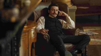 House of Guinness TV show screenshot 1: Anthony Boyle slouches in a brown armchair. He wears a white shirt, black vest, black pants, and black boots.