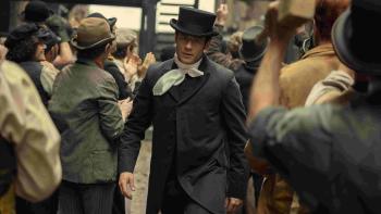 House of Guinness TV show screenshot 2: James Norton walks against a crowd of people in a street. He wears a black top hat and a black coat.