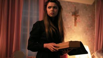 Hysteria! TV show screenshot 2:Fai stands in her room holding open a bible and glancing to the side with furrowed eyebrows. A crucifix is on the wall.