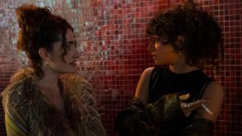 I Love LA TV show screenshot 3: Maia and Tallulah face each other in front of a red tile background; they look to be in serious conversation