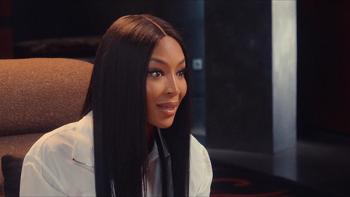 Supermodel Naomi Campbell is pictured sitting in a brown chair, speaking. Her hair is long, dark, and straightened.