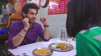 A South Asian man in a plaid shirt talks to a South Asian woman wearing green as they sit at a table in a restaurant.