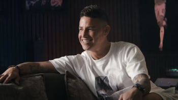 James. screenshot 2: James Rodríguez smiling on a sofa in a dimly lit room, wearing a white T-shirt.
