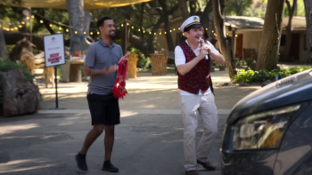 Jury Duty Company Retreat TV show screenshot 1: Steve and Kevin stand in a parking lot together; Kevin is on a microphone and wearing a captain's hat