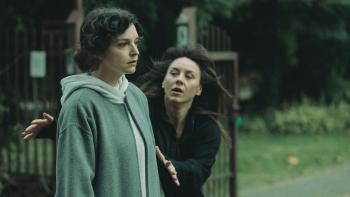 One woman wearing all black rushes to another woman who wears drab grey and looks dazed and exhausted. They are both outside, it looks dark and cold