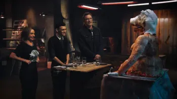 Darkened room with the three show hosts smiling uncertainly and grimacing at a large cake in the shape of a fleshy pig-like being.
