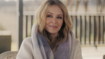 Kylie TV show screenshot 1: Kylie Minogue smiles while seated outdoors wearing a cozy scarf.