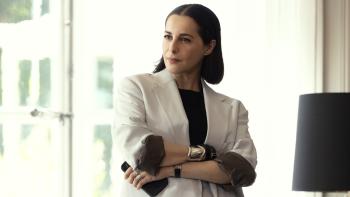 La Maison TV show screenshot 3: Amira Casar wears a white suit jacket, a black shirt, gold bangles, and holds a phone.