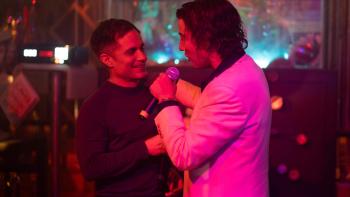 Gael García Bernal and Diego Luna, holding a microphone up, sing karaoke together under the pink lights of a club.