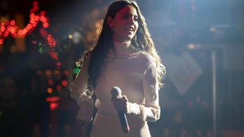Eiza González dances in the club wearing a beige outfit. She is under a spotlight and holds a microphone.