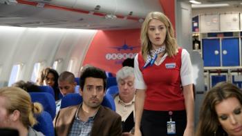 LA to Vegas TV Show: A flight attendant stands next to a passenger; both are looking offscreen and appear to be surprised. 